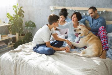 A close-knit family of four, including two parents and two children, sits together on a bed with a large, friendly dog. The parents are seated on either side of the children, with one child on the father's lap and the other next to the mother. The mother lovingly brushes the face of her son while the dog lays beside them, adding to the peaceful and affectionate atmosphere. The room is bright with natural light coming through large windows, with plants in the background, creating a cozy and nurturing environment.