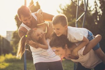 A family of four, including both parents and two children, is having a playful moment outdoors in a park or playground. The father is carrying a child on his back while the mother is playfully lifting another child. Both parents are smiling widely, while the children, who are laughing, seem to enjoy the outdoor fun. The golden hour sunlight casts a soft glow on the family, enhancing the warmth and joy of the scene. The background shows a bright blue sky and green trees, adding to the lively, carefree atmosphere of the moment.