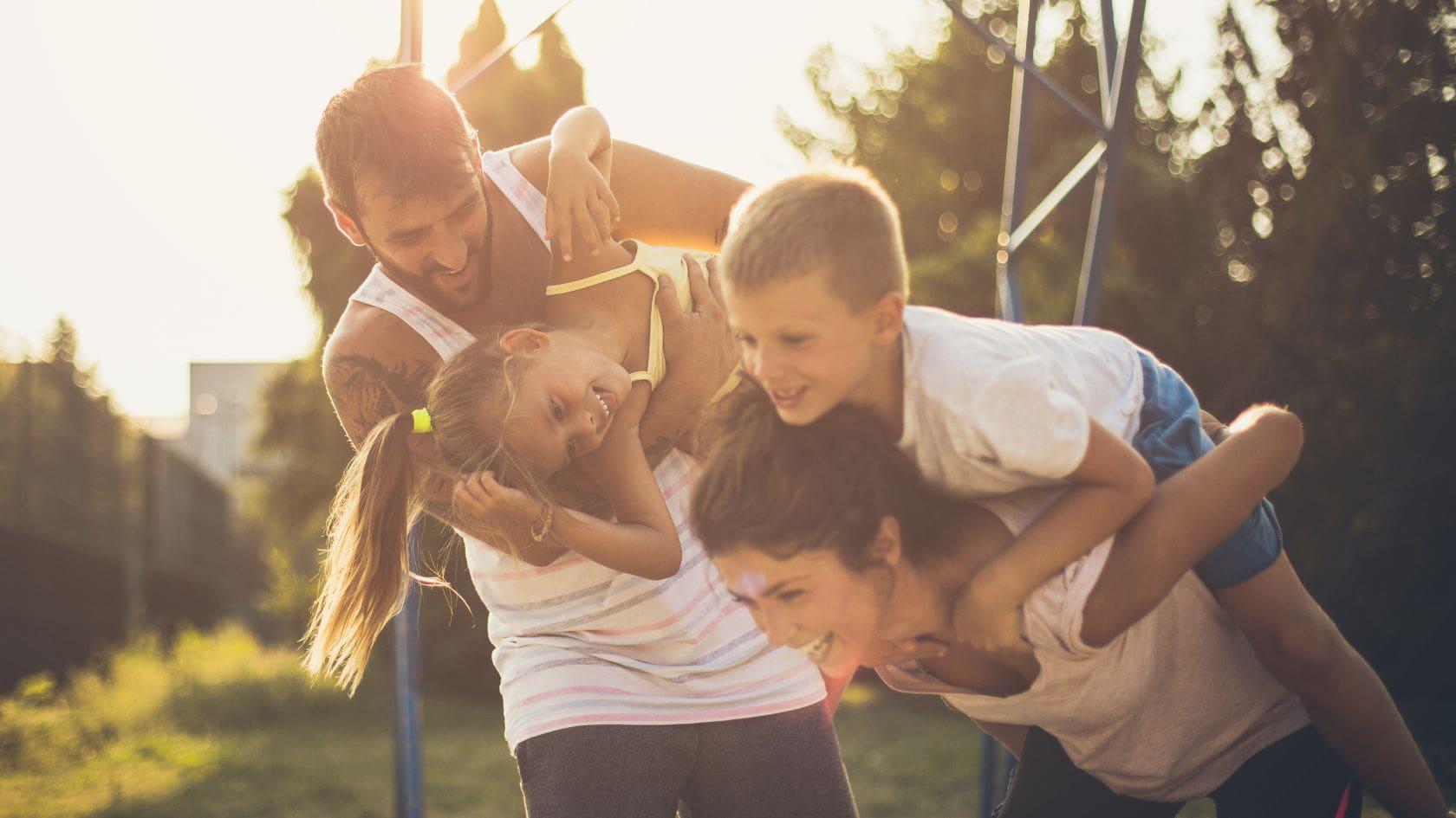 A family of four, including both parents and two children, is having a playful moment outdoors in a park or playground. The father is carrying a child on his back while the mother is playfully lifting another child. Both parents are smiling widely, while the children, who are laughing, seem to enjoy the outdoor fun. The golden hour sunlight casts a soft glow on the family, enhancing the warmth and joy of the scene. The background shows a bright blue sky and green trees, adding to the lively, carefree atmosphere of the moment.