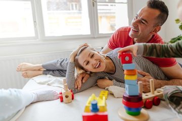 A happy family of three, a mother, father, and young child, is playing together on a bed. The parents are lying on the bed, smiling joyfully as the father gently supports the mother. The child, who is standing nearby, is stacking colorful wooden blocks in front of them. The soft natural light filters through a window, creating a warm and cozy atmosphere. The room is simple and light-filled, with wooden toys scattered around, adding to the playful and loving environment.