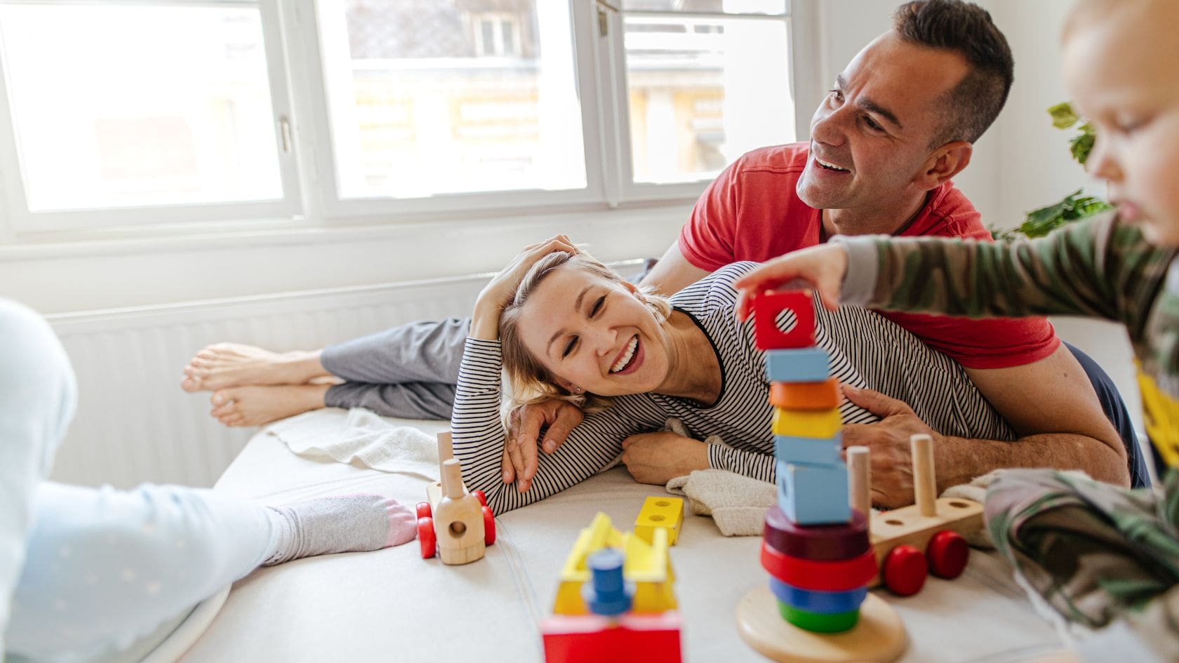 A happy family of three, a mother, father, and young child, is playing together on a bed. The parents are lying on the bed, smiling joyfully as the father gently supports the mother. The child, who is standing nearby, is stacking colorful wooden blocks in front of them. The soft natural light filters through a window, creating a warm and cozy atmosphere. The room is simple and light-filled, with wooden toys scattered around, adding to the playful and loving environment.