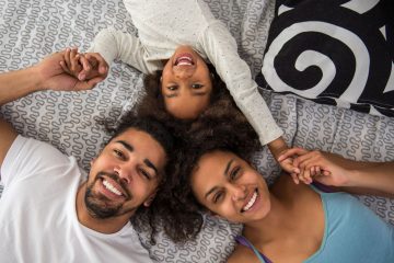 A happy family of three—mom, dad, and a young child—are lying on a bed together, enjoying a fun and relaxed moment. The parents are holding hands with their child, whose face is upside down and laughing joyously. The whole family is smiling, with the parents gazing lovingly at their child, while the child beams with joy. The setting has a calm and relaxed vibe, with a soft white bedspread and decorative pillows, as well as light streaming through the window.