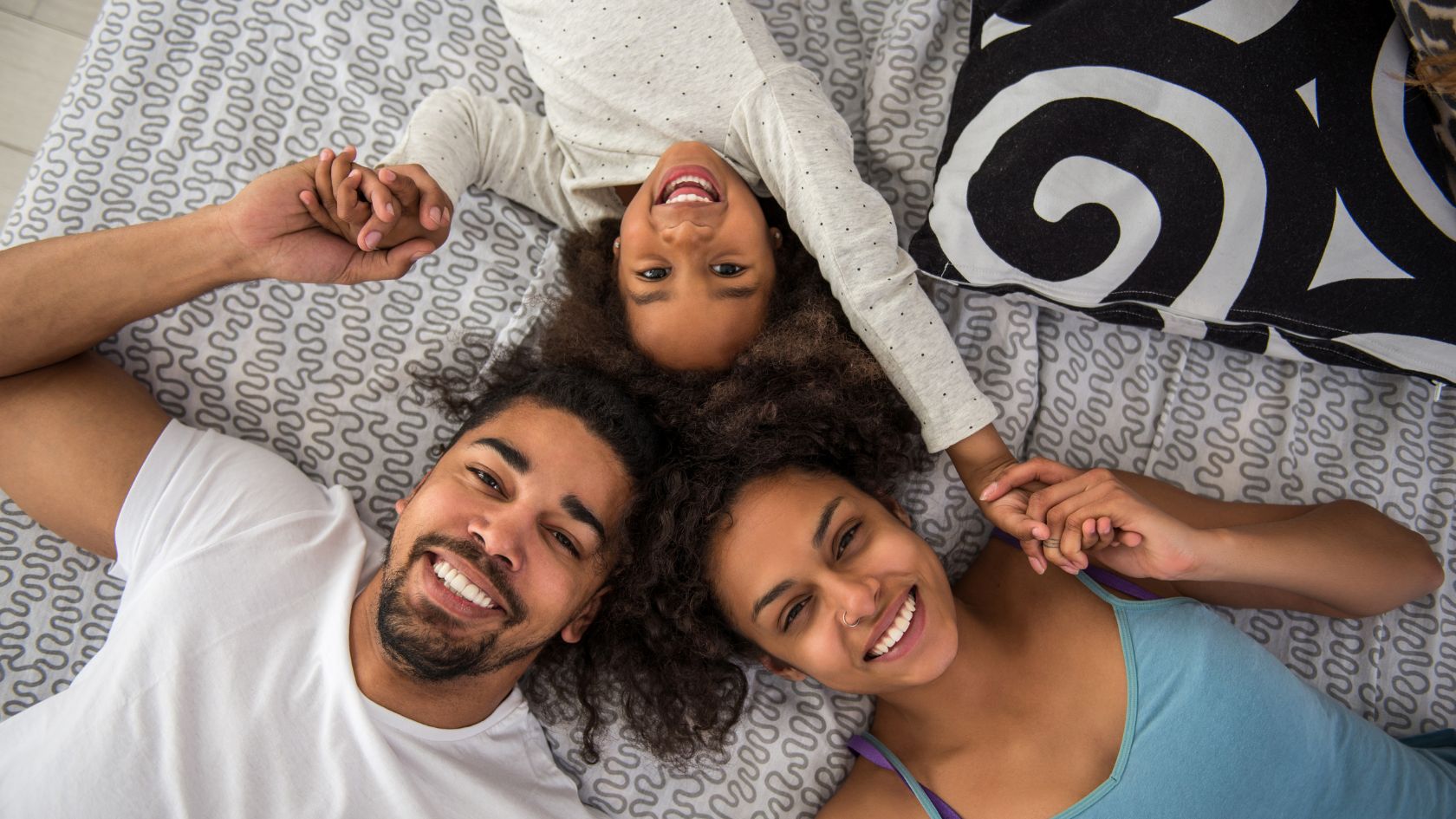 A happy family of three—mom, dad, and a young child—are lying on a bed together, enjoying a fun and relaxed moment. The parents are holding hands with their child, whose face is upside down and laughing joyously. The whole family is smiling, with the parents gazing lovingly at their child, while the child beams with joy. The setting has a calm and relaxed vibe, with a soft white bedspread and decorative pillows, as well as light streaming through the window.