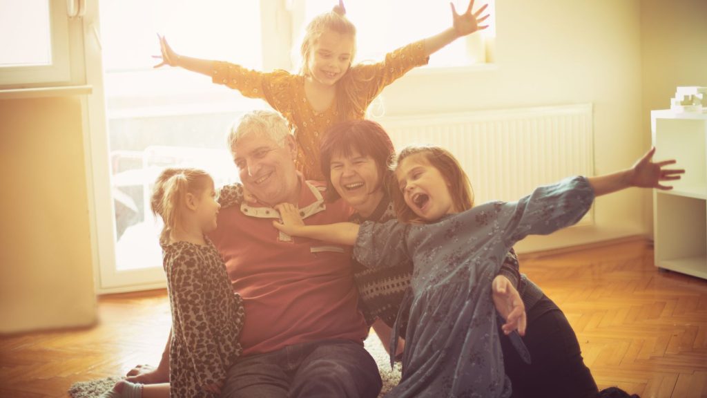 A joyful, multi-generational family enjoys a fun moment together on the floor. In this image, a grandmother and grandfather sit smiling while their three grandchildren, who are full of energy, laugh and play around them. The light from a nearby window creates a warm, golden glow that adds to the cozy atmosphere. The children are in playful poses, with arms outstretched and wide smiles, as the adults share their happiness. The setting is a comfortable living room with soft carpet and furniture.