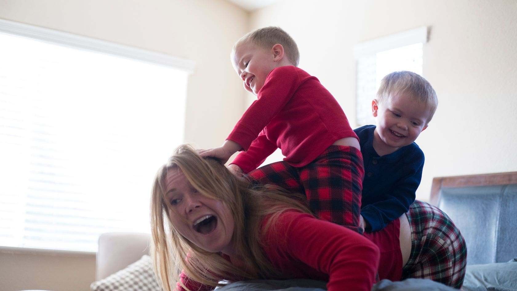 A mother is playfully engaging with her two young children on a bed. The children, dressed in matching red and blue plaid pajamas, are climbing over their mother, with big smiles on their faces as they have fun. The mother, laughing, is lying on her stomach while the boys joyfully sit on her back, creating an amusing and lively moment. The bright light from the window enhances the cheerful and energetic mood of the scene, reflecting a playful family interaction.