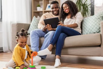 Family Bonding at Home – A young couple sits on a cozy couch, smiling as they look at a tablet, while their toddler happily plays on the floor, showcasing love, technology, and connection