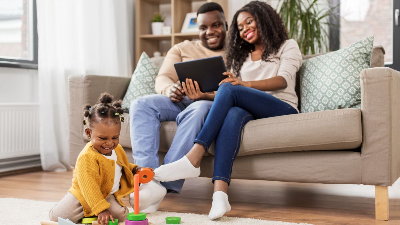 Family Bonding at Home – A young couple sits on a cozy couch, smiling as they look at a tablet, while their toddler happily plays on the floor, showcasing love, technology, and connection