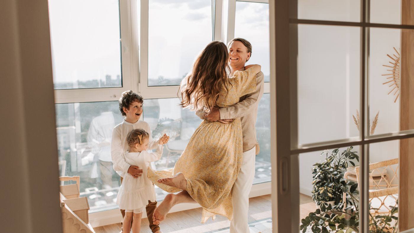 Joyful Homecoming – A mother leaps into her partner’s arms as their children excitedly watch, illustrating the happiness of returning home and cherishing family moments